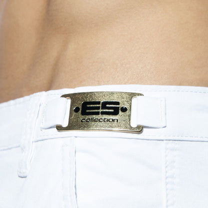 Close-up of white ES Collection shorts featuring a gold-toned metal logo plaque on the waistband.