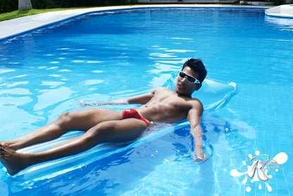 A man wearing icker sea lifeguard micro slip bikini swimwear red floats on a translucent blue inflatable pool float