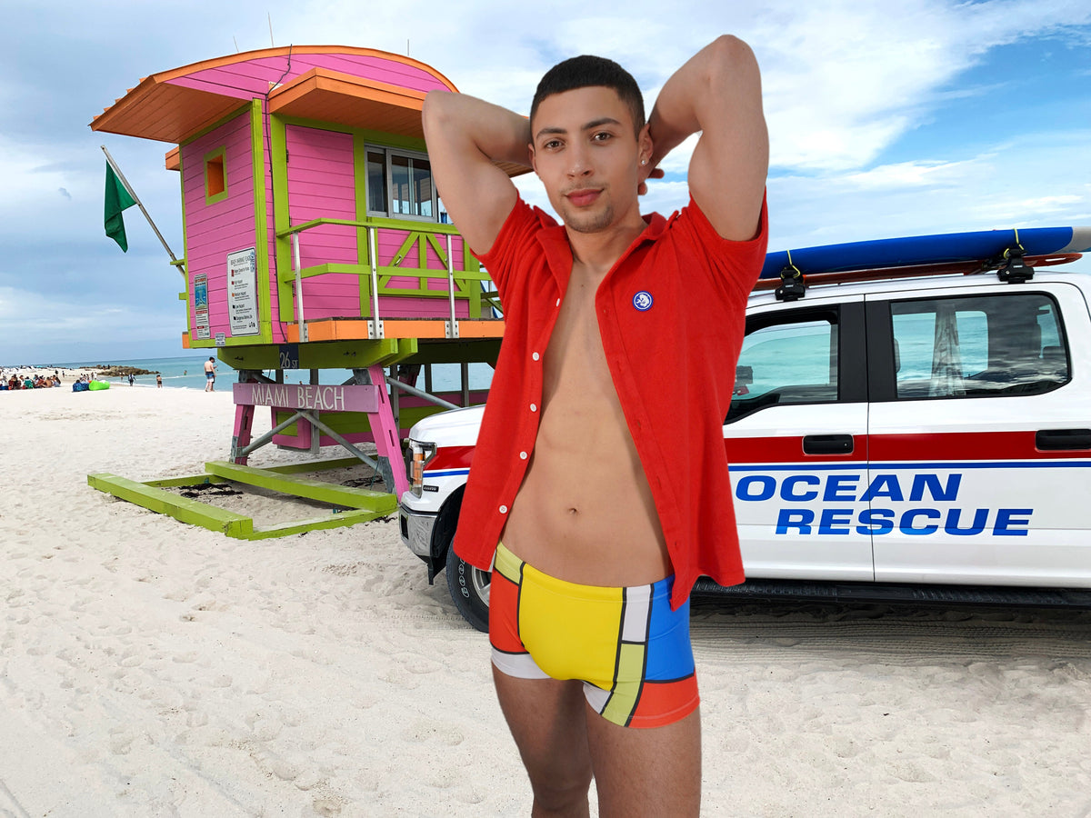 Colorful square cut men's swimsuit by Maris Equi featuring a vibrant geometric pattern worn by a model at the beach.