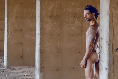 A muscular man with long dark hair and tattoos wears a turquoise JUSTIN+SIMON XSJ02 classic g-string bulge against graffiti