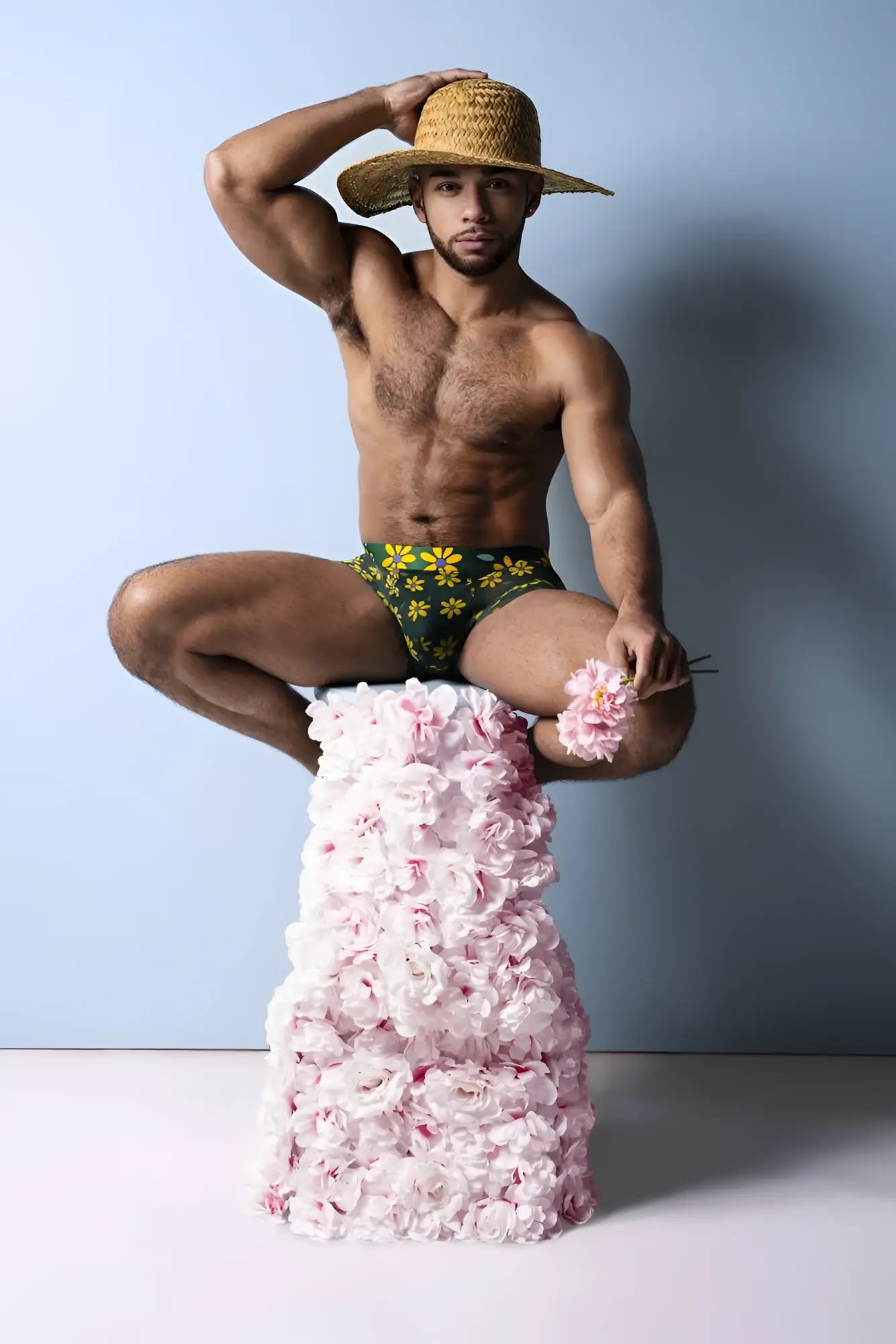 A man wearing a straw hat and male power petal power pouch shorts with green daisy print sits on pink roses pedestal