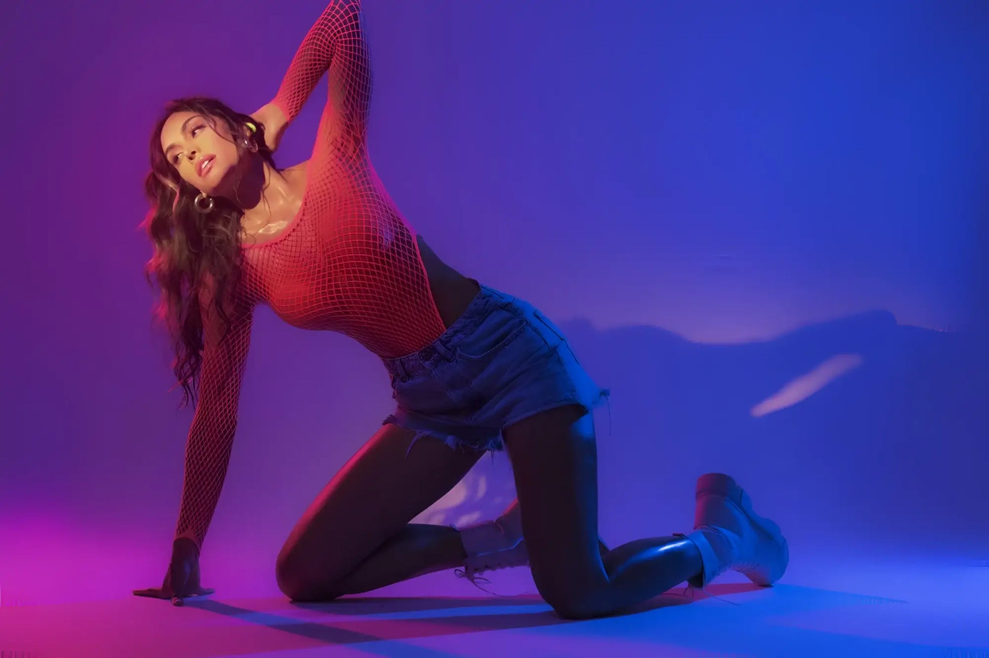 A woman poses in a neon pink Mapale 1119 long sleeve fishnet bodysuit with denim shorts under purple pink lighting