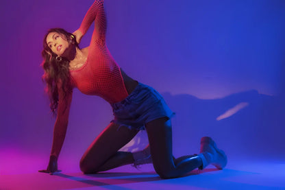 A woman poses in a neon pink Mapale 1119 long sleeve fishnet bodysuit with denim shorts under purple pink lighting