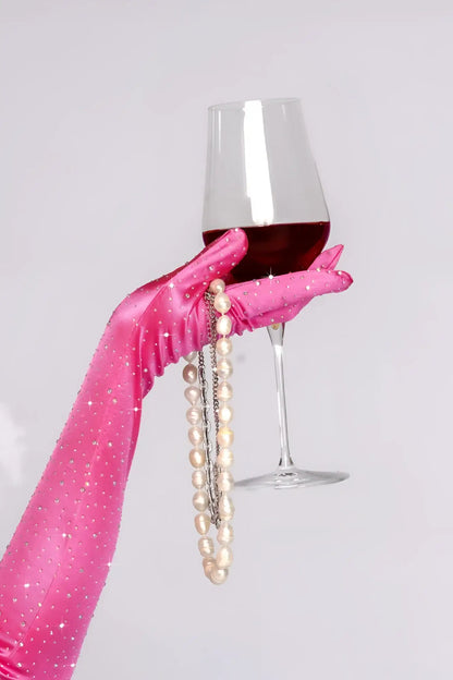 A hand in mapale 135 pink satin rhinestone opera gloves holds a red wine glass with pearl necklaces