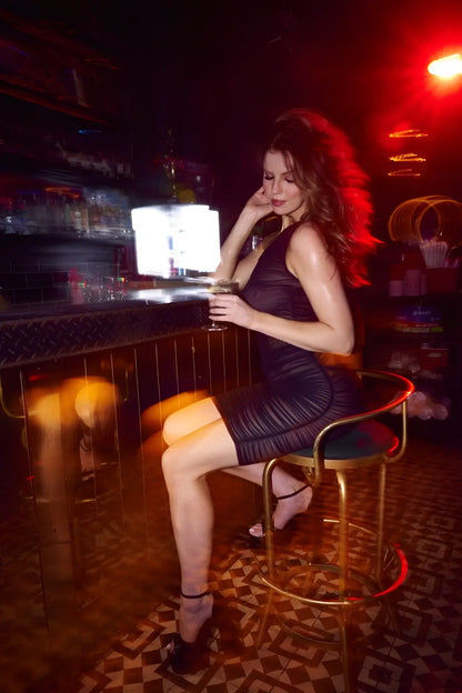 A woman wearing a form-fitting black Mapale 40002 dress sits on a gold bar stool holding a glass of wine
