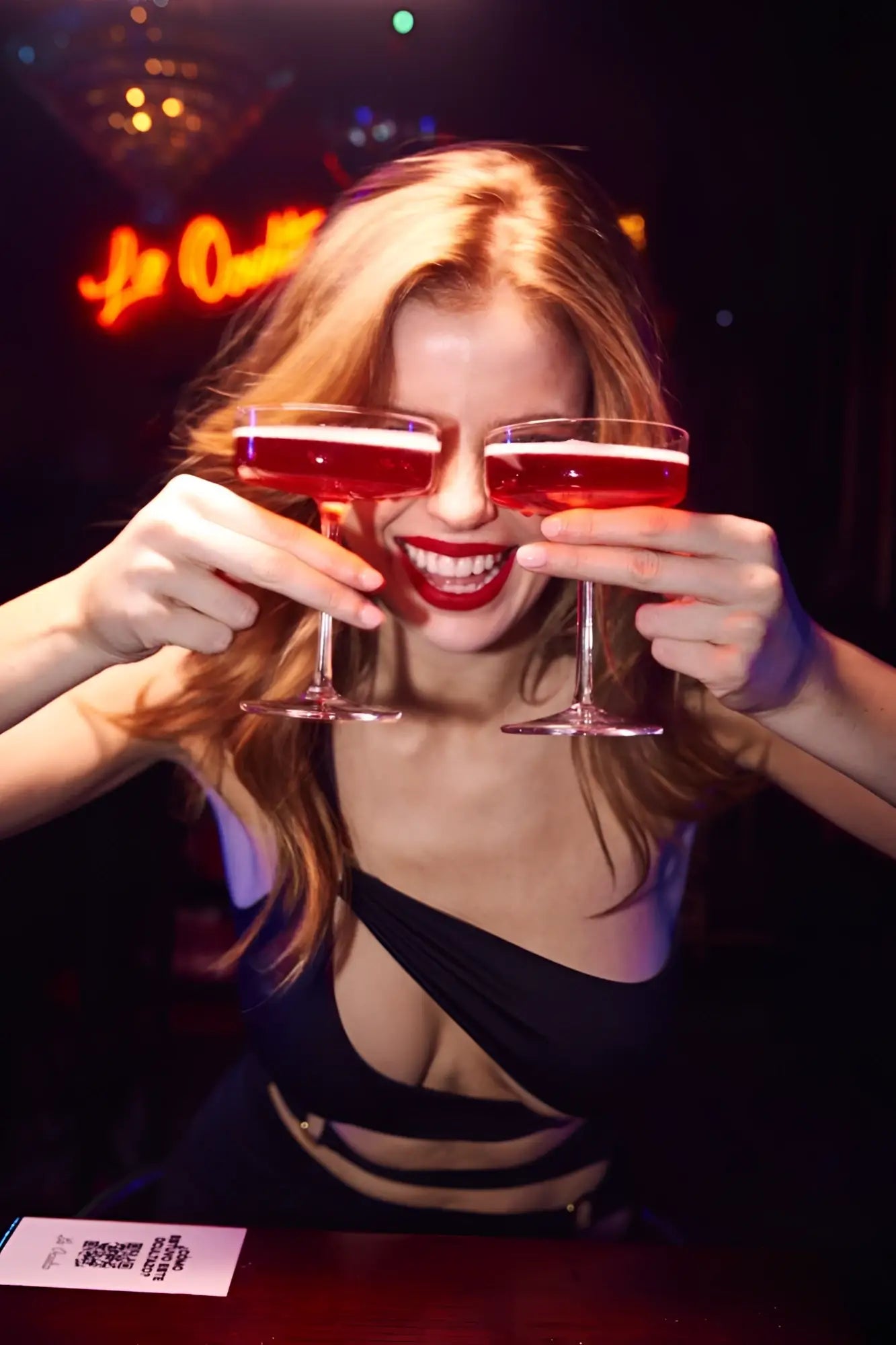 A woman with blonde hair holding two coupe glasses stands wearing a black mapale 40006 dress
