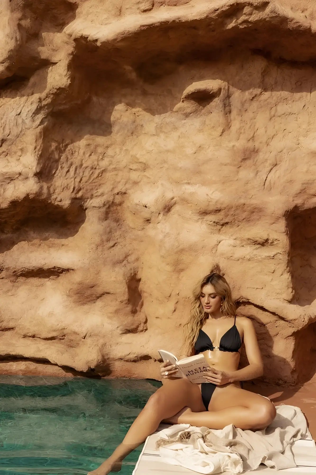 A woman sits by the poolside wearing a black Mapale 67166 two piece swimsuit reading a book titled milia