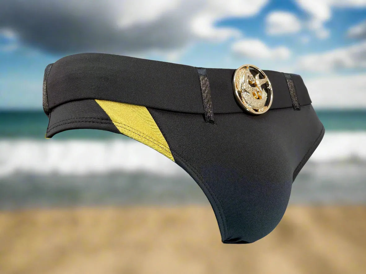 Black swim briefs with yellow side panel and gold medallion detail by Maris Equi shown against a beach background.
