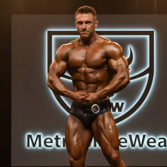A muscular male bodybuilder wearing black shiny crocodile swim briefs with a silver number 3 badge on the waistband