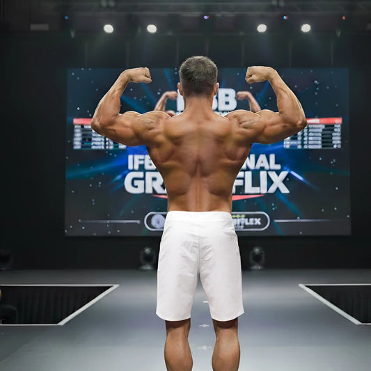 A muscular man wearing metro malewear physique board short white 4706-00 stands on stage showing back side