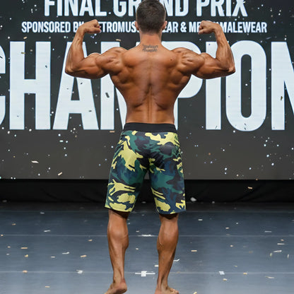 A muscular man stands on stage wearing MetroMaleWear physique board shorts in yellow green camo pattern