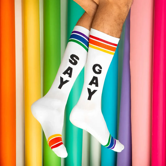 White crew socks with black say gay lettering and rainbow striped cuffs toe caps and heels in knee high style