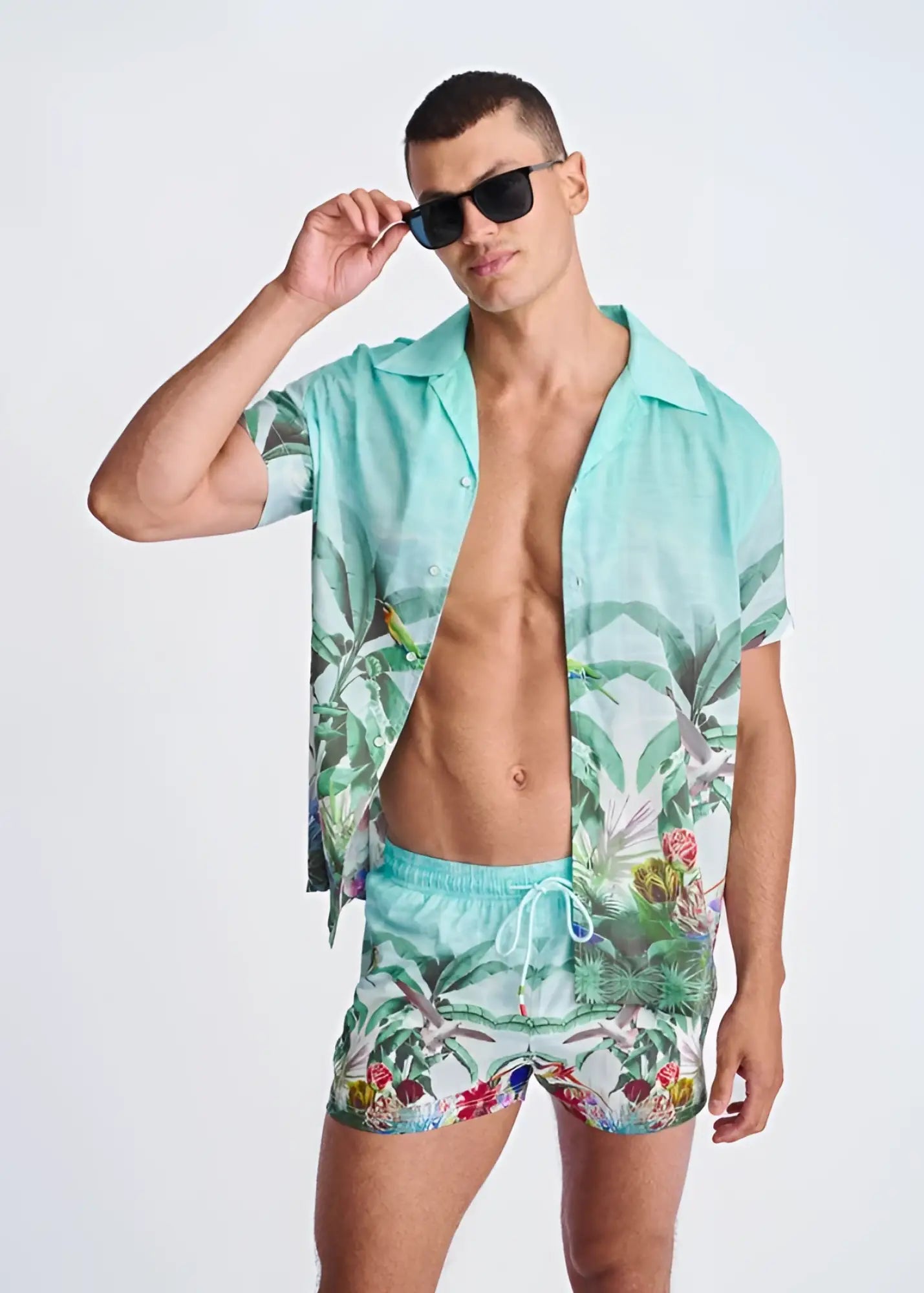 A vibrant turquoise tropics print swim shorts with mesh lining featuring palm leaves and birds paired with matching shirt