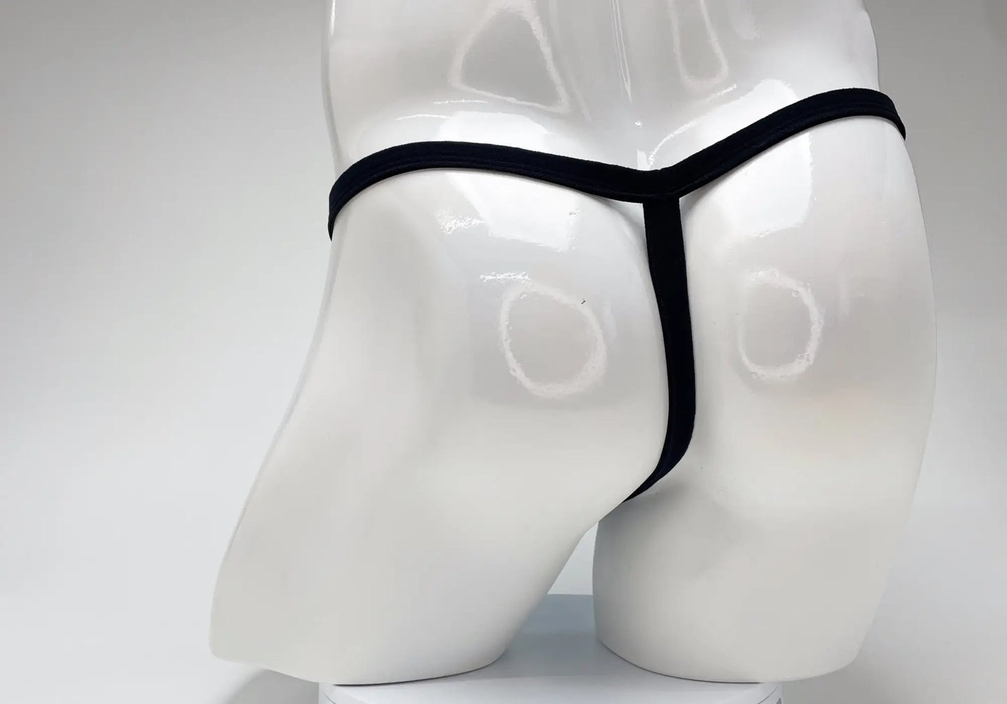 Back view of a black thin-strap thong on a glossy white male mannequin torso.