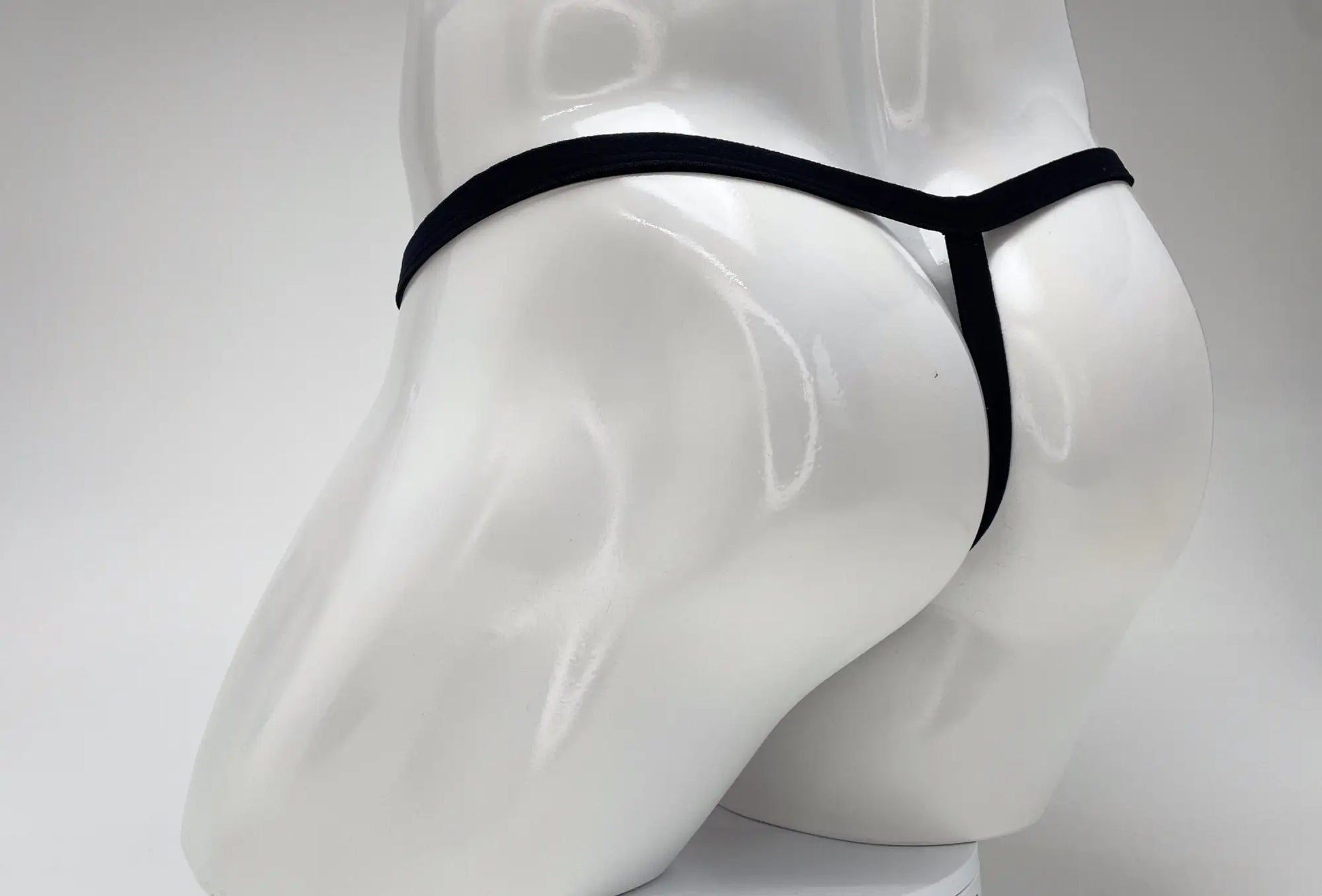 Back view of a white male mannequin wearing a minimalist black thong with a thin waistband and vertical strap.