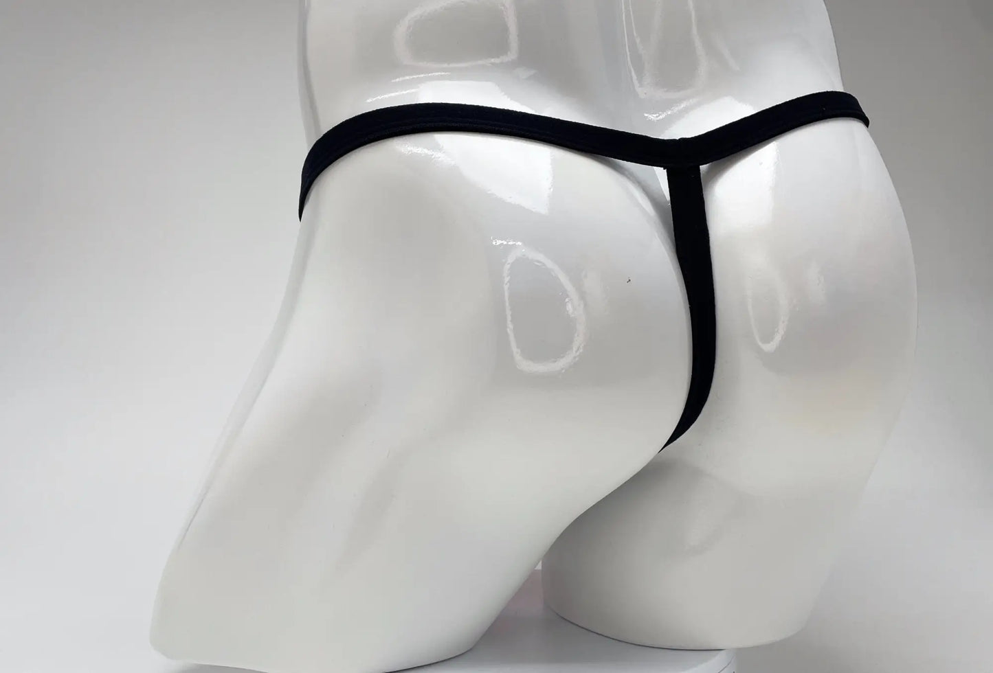 Rear view of a white male mannequin wearing a minimal black thong with a thin waistband and vertical strap.